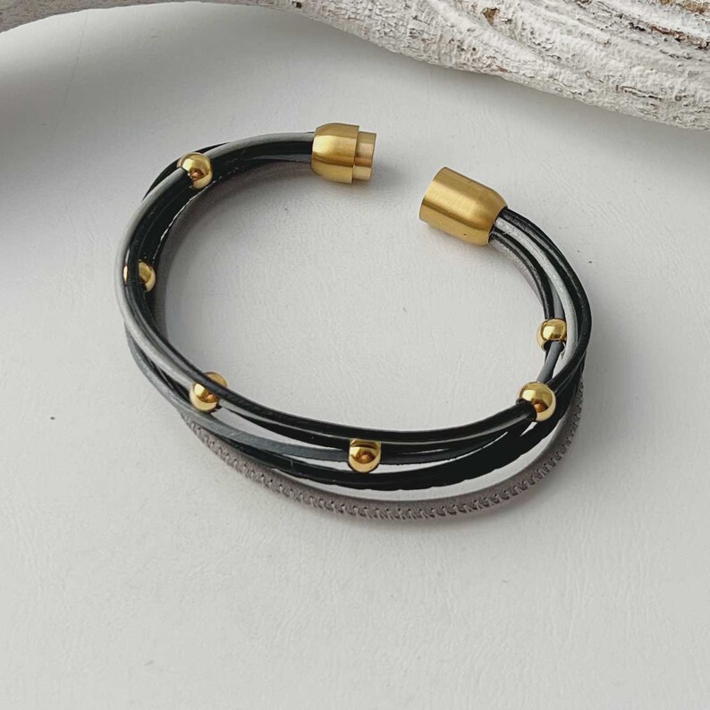 K&ouml;nnte beinhalten: Ein mehrreihiges Lederarmband mit einem Magnetverschluss. Das Armband besteht aus schwarzem, wei&szlig;em und grauem Leder und hat goldfarbene Perlen.