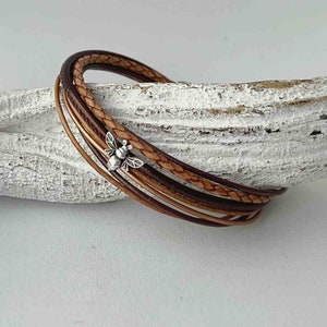 May include: A multi-strand leather bracelet with a braided brown leather strap and a plain brown leather strap. A small silver bee charm is attached to the bracelet.