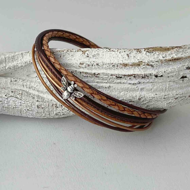 May include: A multi-strand leather bracelet with a braided brown leather strap and a plain brown leather strap. A small silver bee charm is attached to the bracelet.