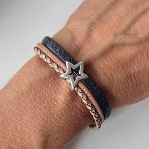 May include: A layered leather bracelet with a star charm. The bracelet features two black leather bands with the words "Every Moment" and "Love Life" embossed on them. The third band is a braided light brown leather band.