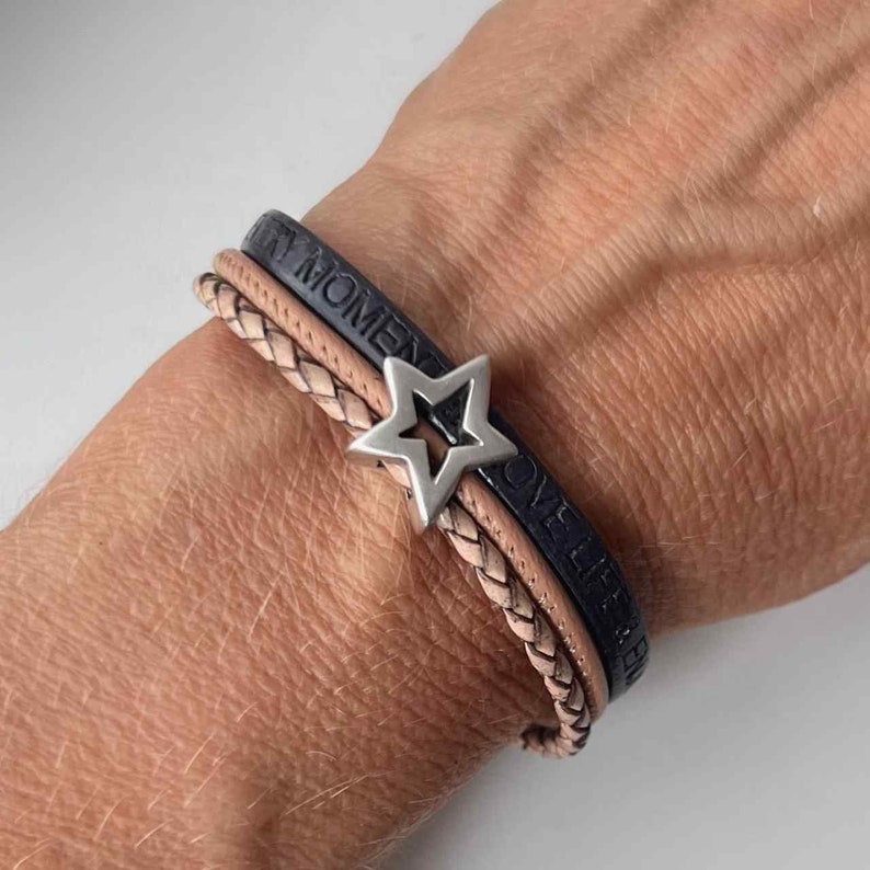 May include: A layered leather bracelet with a star charm. The bracelet features two black leather bands with the words "Every Moment" and "Love Life" embossed on them. The third band is a braided light brown leather band.
