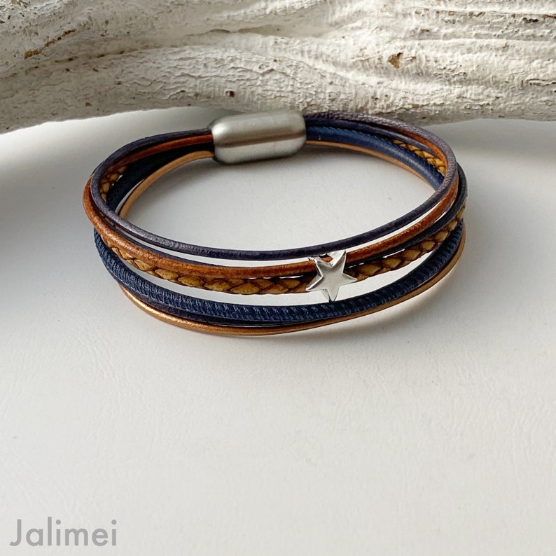 May include: A multi-layered leather bracelet with a silver star charm. The bracelet is made of brown and blue leather and has a silver metal clasp.