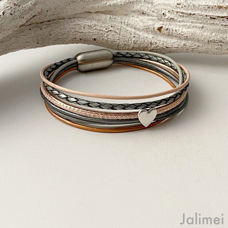 K&ouml;nnte beinhalten: Ein mehrstr&auml;ngiges Armband mit Magnetverschluss. Das Armband enth&auml;lt Str&auml;nge in verschiedenen Farben, darunter Grau, Rosa und Kupfer, mit einem silbernen Herzanh&auml;nger. Das Design umfasst geflochtene und glatte Lederstrukturen.