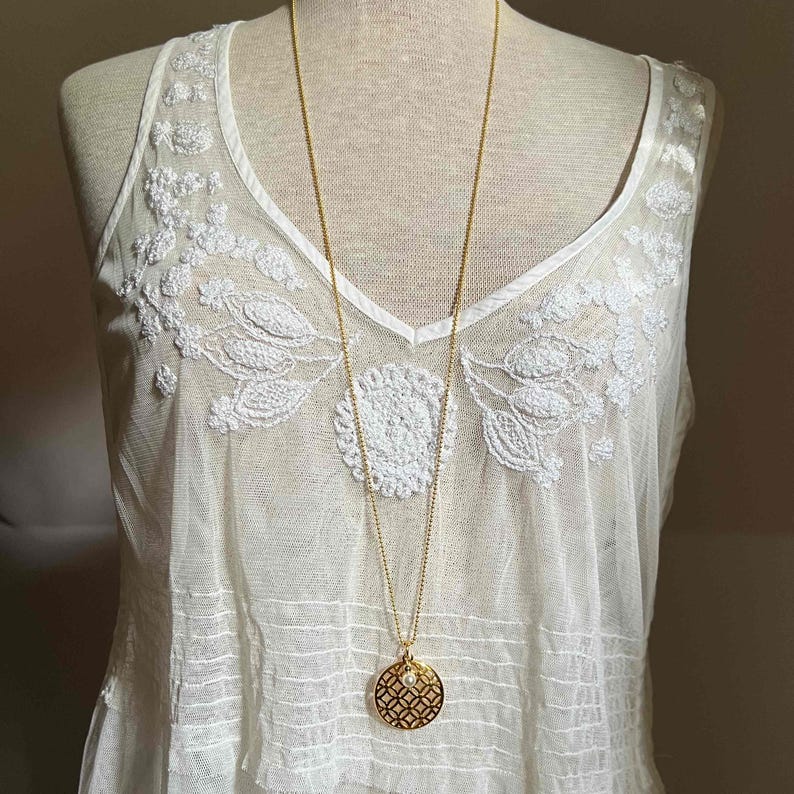 Puede incluir: Un collar largo dorado con un colgante circular con un patr&oacute;n geom&eacute;trico y una peque&ntilde;a perla. El collar cuelga sobre una camiseta sin mangas blanca bordada en un maniqu&iacute;.