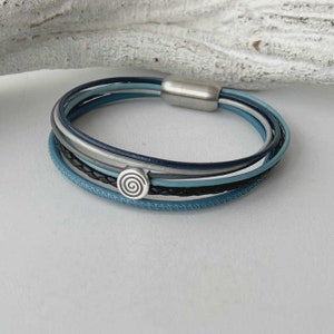 May include: Multi-layered leather bracelet with a silver clasp and a silver spiral charm. The bracelet is made of blue, gray, and black leather.