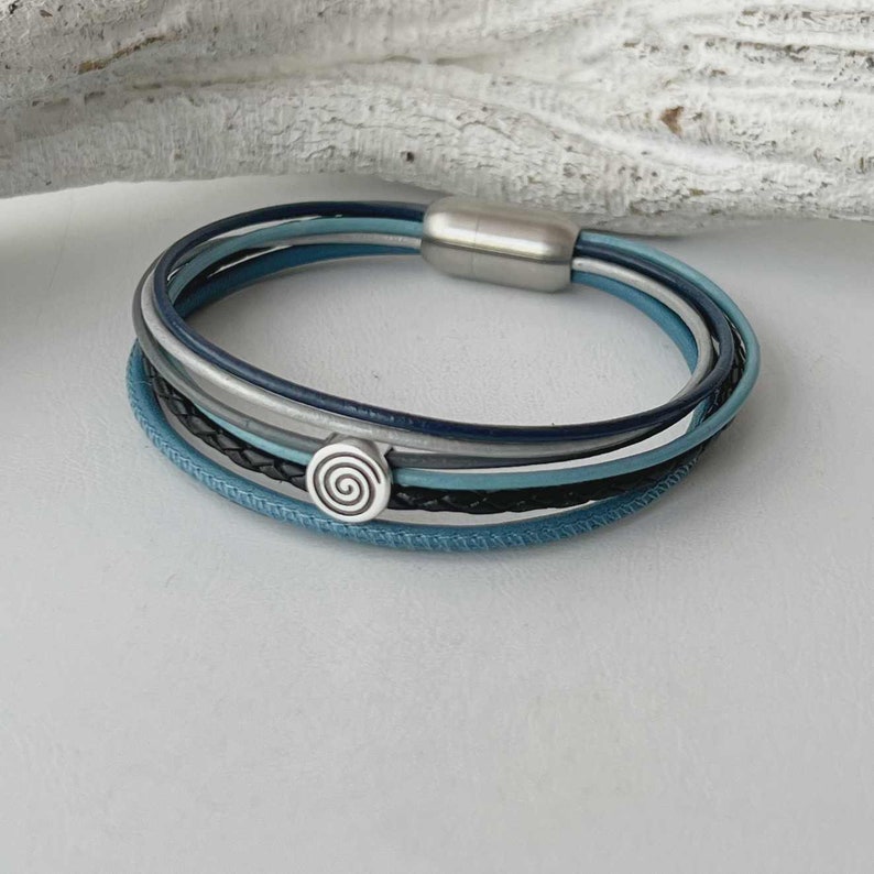 May include: Multi-layered leather bracelet with a silver clasp and a silver spiral charm. The bracelet is made of blue, gray, and black leather.