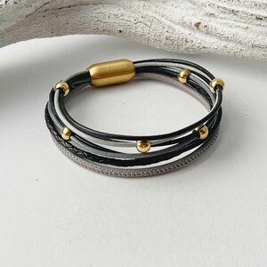 K&ouml;nnte beinhalten: Ein mehrreihiges Lederarmband mit einem Magnetverschluss. Das Armband besteht aus schwarzen, grauen und wei&szlig;en Lederstr&auml;ngen mit goldenen Perlen.