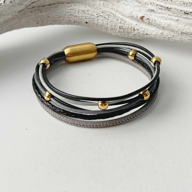 K&ouml;nnte beinhalten: Ein mehrreihiges Lederarmband mit einem Magnetverschluss. Das Armband besteht aus schwarzen, grauen und wei&szlig;en Lederstr&auml;ngen mit goldenen Perlen.
