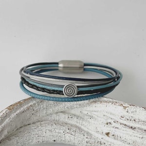 May include: A multi-layered bracelet with a silver magnetic clasp and a silver spiral charm. The bracelet is made of blue, gray, and black leather.