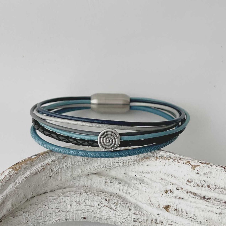 May include: A multi-layered bracelet with a silver magnetic clasp and a silver spiral charm. The bracelet is made of blue, gray, and black leather.