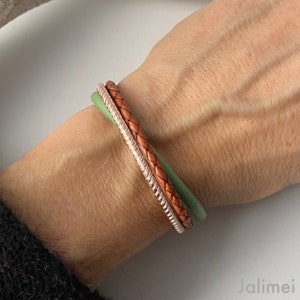May include: A stack of three bracelets in shades of brown, silver, and green. The brown bracelet has a braided design, the silver one has a textured surface, and the green one is smooth.