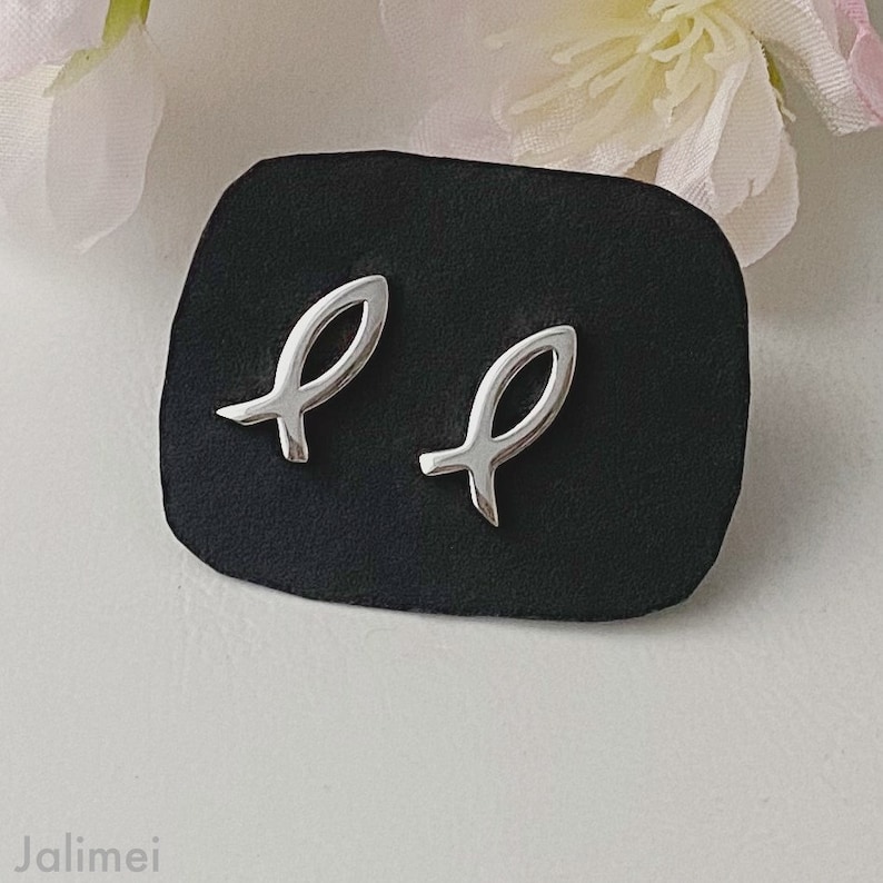 May include: A pair of silver fish-shaped earrings on a black velvet display. The earrings are small and delicate.