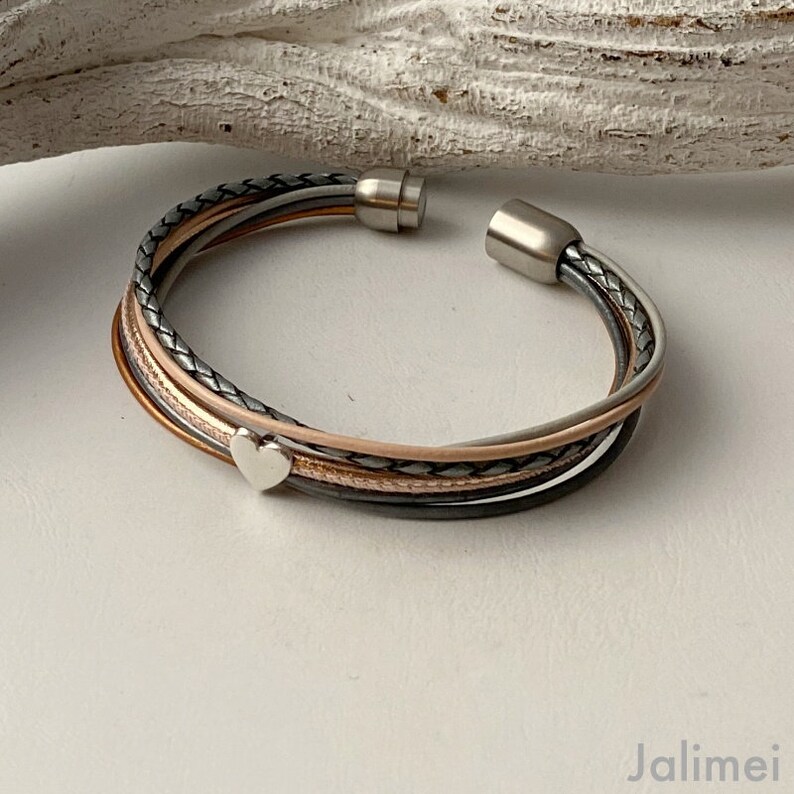 K&ouml;nnte beinhalten: Ein mehrstr&auml;ngiges Armband mit Magnetverschluss. Das Armband hat ineinander verschlungene Str&auml;nge in Grau-, Schwarz-, Kupfer- und Rosat&ouml;nen. Ein kleiner silberner Herzanh&auml;nger ist befestigt. Das Armband ist offen und zeigt den Verschluss.