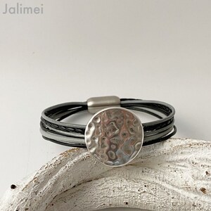 K&ouml;nnte beinhalten: Ein mehrreihiges Lederarmband mit einem silbernen, geh&auml;mmerten, runden Anh&auml;nger. Das Armband besteht aus schwarzem, grauem und wei&szlig;em Leder.