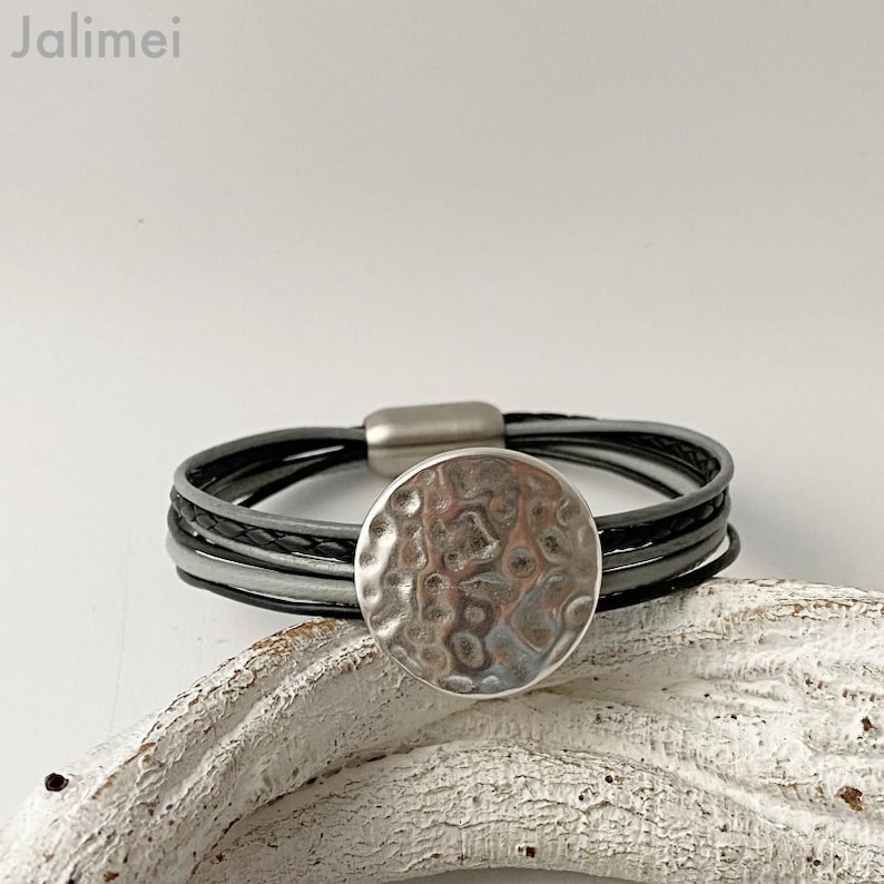 K&ouml;nnte beinhalten: Ein mehrreihiges Lederarmband mit einem silbernen, geh&auml;mmerten, runden Anh&auml;nger. Das Armband besteht aus schwarzem, grauem und wei&szlig;em Leder.