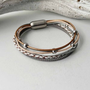 May include: A multi-strand bracelet with a magnetic clasp. The bracelet is made of braided leather in shades of grey, white, and pink. The bracelet is accented with silver beads.