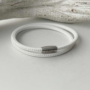 Könnte beinhalten: Ein weißes Lederarmband mit Wickeldesign. Das Armband hat eine genähte Textur und einen silberfarbenen Verschluss mit dem eingravierten Wort "Jaimei". Das Armband wird auf einer weißen Oberfläche gezeigt.