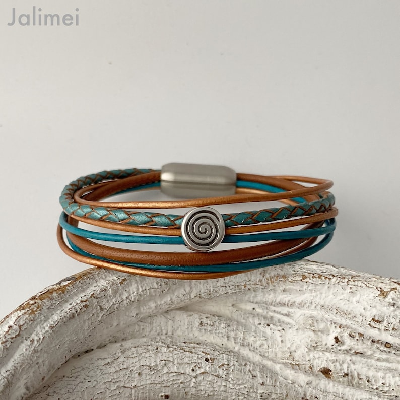 Ein mehrlagiges Lederarmband mit einem silbernen Spiral-Anhänger. Das Armband besteht aus braunem und türkisfarbenem Leder und hat einen Magnetverschluss.