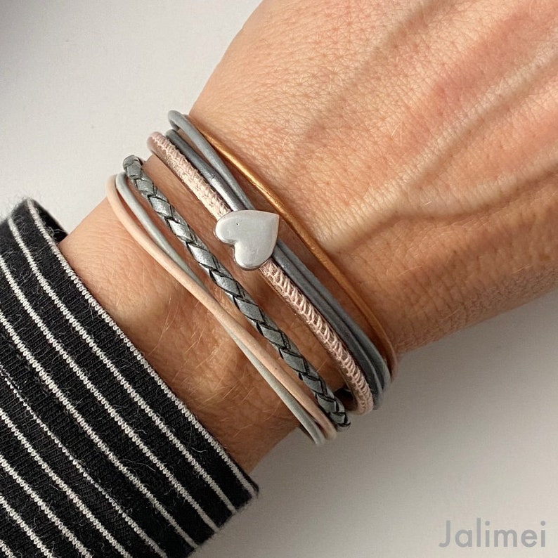 K&ouml;nnte beinhalten: Ein mehrstr&auml;ngiges Armband mit einem silbernen Herzanh&auml;nger. Das Armband enth&auml;lt Str&auml;nge in verschiedenen Farben: Hellrosa, Grau, silber geflochten und einen goldfarbenen Strang. Das Armband wird an einem Handgelenk gezeigt, mit einem schwarz-wei&szlig; gestreiften &Auml;rmel sichtbar.