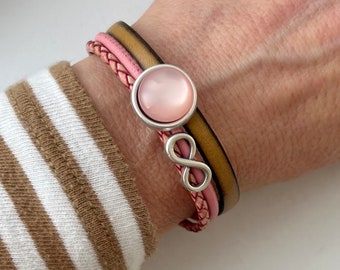 May include: A pink and brown leather bracelet with a silver infinity charm and a pink glass cabochon. The bracelet is on a person's wrist.