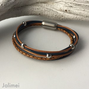 K&ouml;nnte beinhalten: Ein mehrreihiges Lederarmband mit einem geflochtenen braunen Lederband und einem blauen Lederband. Das Armband hat silberne Metallperlen und einen Magnetverschluss.