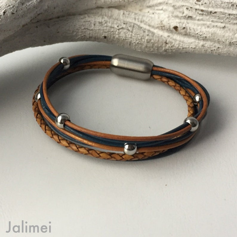 K&ouml;nnte beinhalten: Ein mehrreihiges Lederarmband mit einem geflochtenen braunen Lederband und einem blauen Lederband. Das Armband hat silberne Metallperlen und einen Magnetverschluss.
