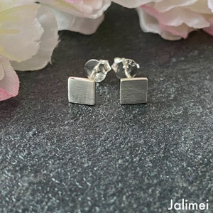 May include: A pair of square silver stud earrings. Each earring has a brushed metal finish and a post backing. The earrings are displayed on a dark gray surface with a blurred floral background.