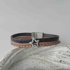 May include: A multi-layered leather bracelet with a silver star charm. The bracelet features the words "Love Life & Enjoy Every Moment" embossed on the leather.