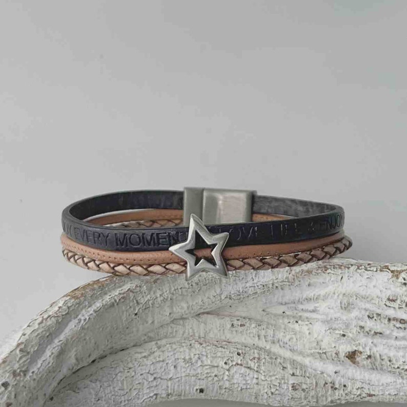 May include: A multi-layered leather bracelet with a silver star charm. The bracelet features the words "Love Life & Enjoy Every Moment" embossed on the leather.