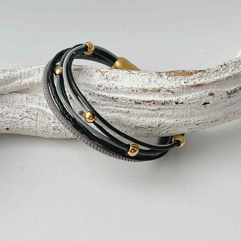 K&ouml;nnte beinhalten: Ein mehrreihiges Armband aus schwarzem, grauem und wei&szlig;em Leder mit goldenen Perlen.