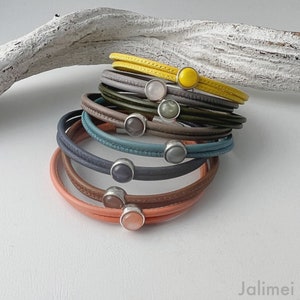 Op de afbeelding: Een stapel leren wikkelarmbanden in verschillende kleuren, waaronder geel, grijs, groen, blauw en koraal. Elke armband heeft een zilveren sluiting met een edelsteen. De armbanden zijn op een wit oppervlak gestapeld.