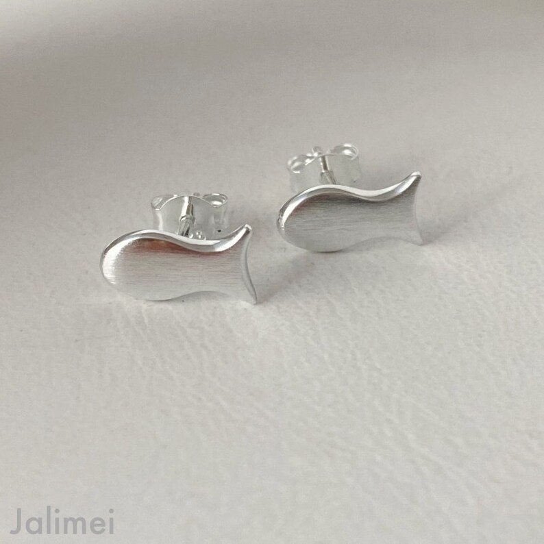 K&ouml;nnte beinhalten: Ein Paar silberne Ohrstecker in Fischform. Die Ohrringe sind klein und zart, mit einer geb&uuml;rsteten Oberfl&auml;che.