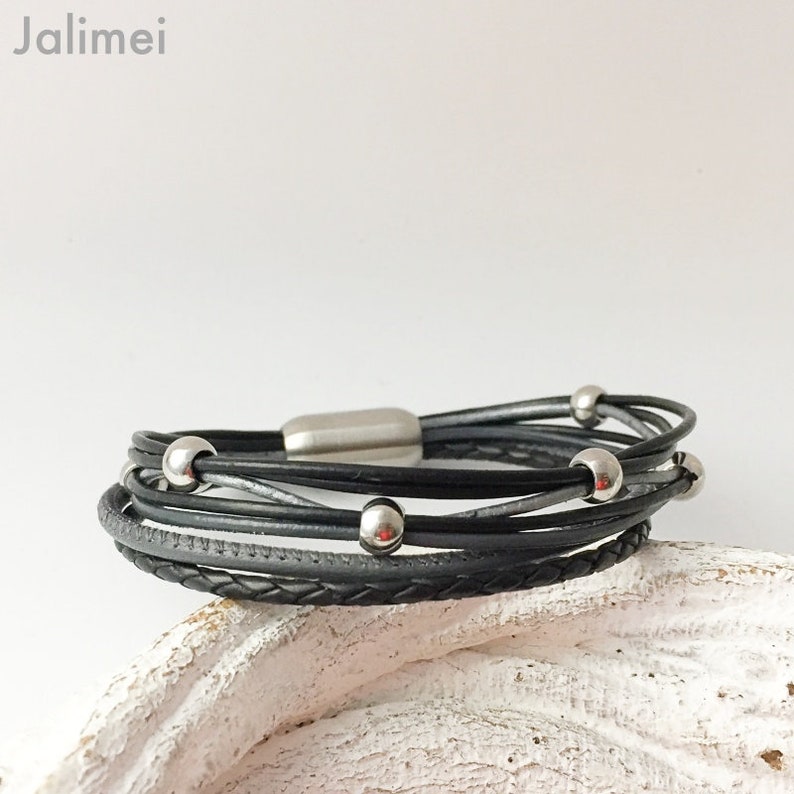 Puede incluir: Un brazalete de cuero negro con una correa de cuero trenzada y cuentas de metal plateado. El brazalete tiene un cierre magn&eacute;tico.
