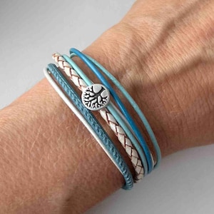 May include: A multi-layered bracelet with a braided leather strap and several thin blue leather straps. A silver charm with a tree design is attached to the bracelet.