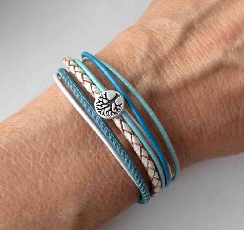 May include: A multi-layered bracelet with a braided leather strap and several thin blue leather straps. A silver charm with a tree design is attached to the bracelet.