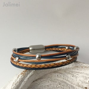 K&ouml;nnte beinhalten: Ein mehrstr&auml;ngiges Lederarmband mit einem geflochtenen braunen Lederband und blauen Lederb&auml;ndern. Das Armband hat silberne Metallperlen und einen Magnetverschluss.