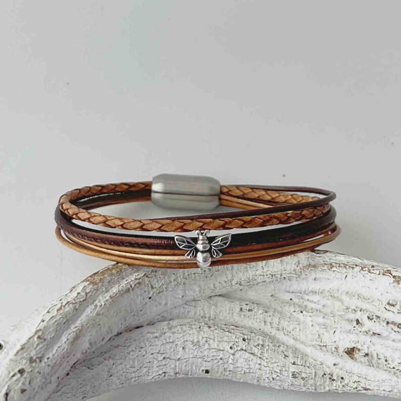 May include: A multi-strand leather bracelet with a silver bee charm. The bracelet is made of brown and tan leather and has a magnetic clasp.