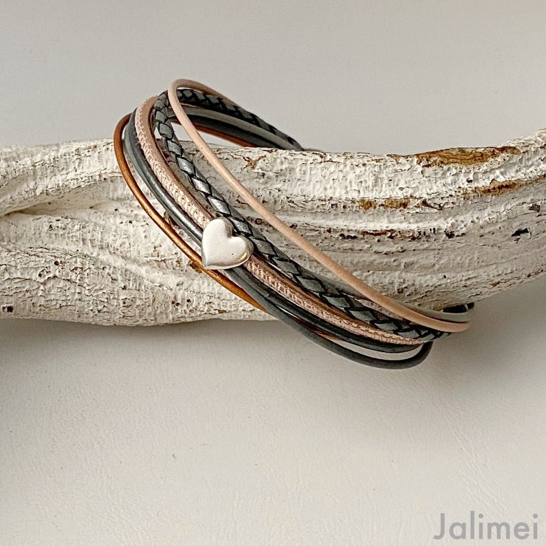 K&ouml;nnte beinhalten: Mehrstrang-Armband mit einem silbernen Herzanh&auml;nger. Das Armband enth&auml;lt Lederstr&auml;nge in Braun, Schwarz, Grau und Rosa. Das Armband im Wickelstil ist so konzipiert, dass es mehrmals um das Handgelenk getragen werden kann.