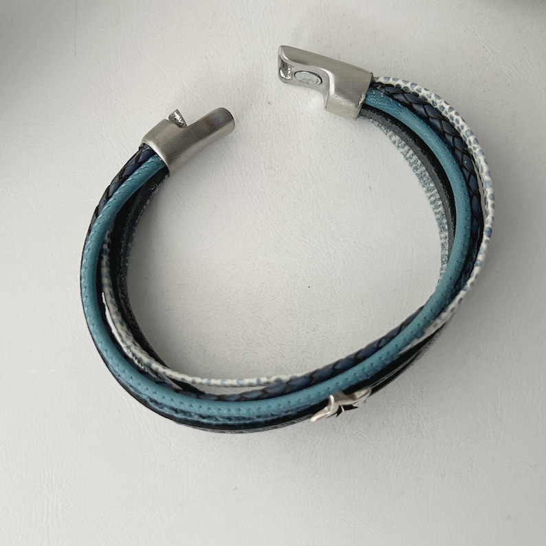 K&ouml;nnte beinhalten: Ein mehrlagiges Lederarmband mit einem silbernen Verschluss. Das Armband besteht aus blauem, schwarzem und wei&szlig;em Leder.