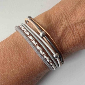 May include: A stack of bracelets featuring a braided leather strap, a grey leather strap, and several thin leather straps with silver beads. The bracelets are worn on a person's wrist.