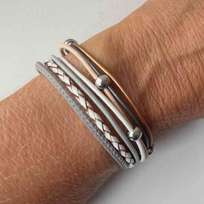 May include: A stack of bracelets featuring a braided leather strap, a grey leather strap, and several thin leather straps with silver beads. The bracelets are worn on a person's wrist.