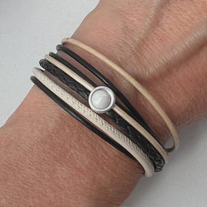 Könnte beinhalten: Ein mehrsträngiges Armband mit schwarzen, cremefarbenen und beigefarbenen Lederkordeln. Es wird mit einem silberfarbenen Magnetverschluss mit einem runden, weißen Stein verschlossen. Das Armband wird um ein Handgelenk getragen.