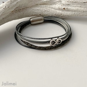 Puede incluir: Un brazalete de cuero multicapa con un dije de infinito plateado. El brazalete est&aacute; hecho de cuero gris oscuro y gris claro.