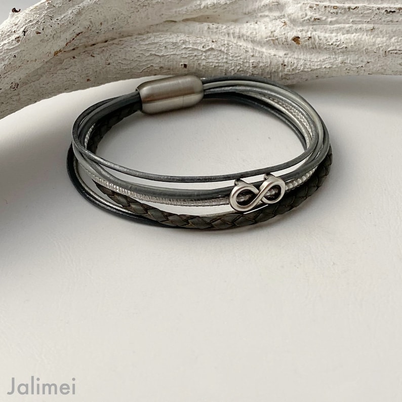 Puede incluir: Un brazalete de cuero multicapa con un dije de infinito plateado. El brazalete est&aacute; hecho de cuero gris oscuro y gris claro.