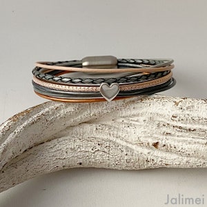 K&ouml;nnte beinhalten: Ein mehrstr&auml;ngiges Armband mit Magnetverschluss. Das Armband hat Str&auml;nge in verschiedenen Farben, darunter Grau, Rosa und Schwarz, mit einem kleinen silbernen Herzanh&auml;nger. Das Armband liegt auf einer wei&szlig;en, strukturierten Oberfl&auml;che.