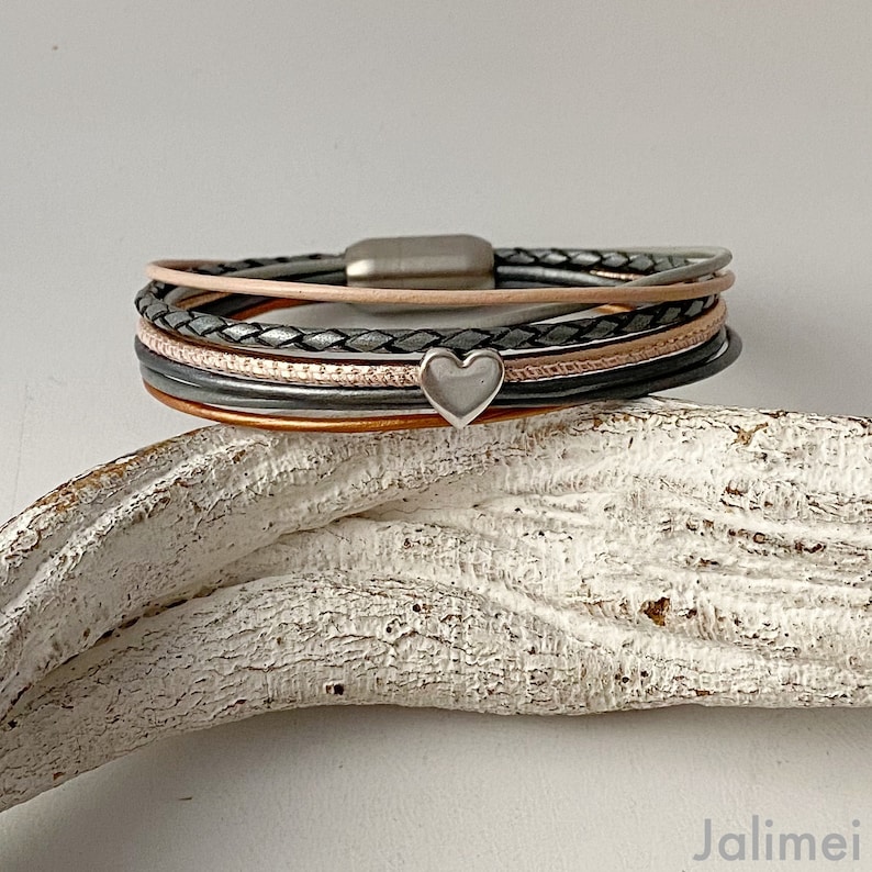 K&ouml;nnte beinhalten: Ein mehrstr&auml;ngiges Armband mit Magnetverschluss. Das Armband hat Str&auml;nge in verschiedenen Farben, darunter Grau, Rosa und Schwarz, mit einem kleinen silbernen Herzanh&auml;nger. Das Armband liegt auf einer wei&szlig;en, strukturierten Oberfl&auml;che.
