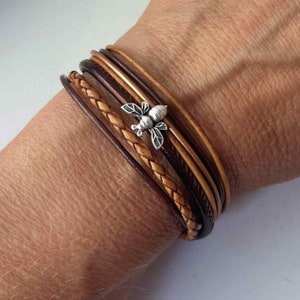 May include: A multi-layered leather bracelet with a braided brown leather strap and a silver bee charm.