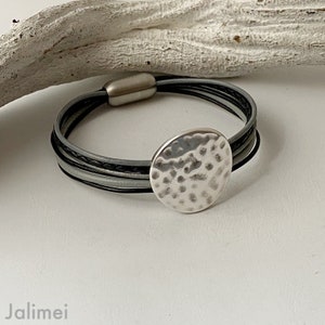 K&ouml;nnte beinhalten: Ein silbernes Armband mit einem geh&auml;mmerten Metallkreis-Anh&auml;nger. Das Armband besteht aus schwarzen und grauen Lederriemen.