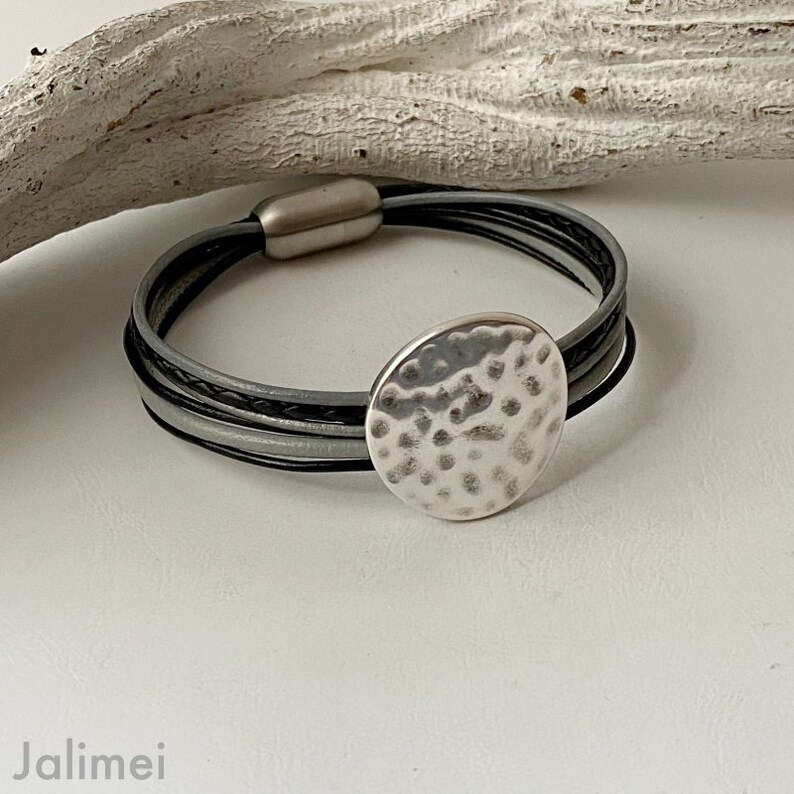 K&ouml;nnte beinhalten: Ein silbernes Armband mit einem geh&auml;mmerten Metallkreis-Anh&auml;nger. Das Armband besteht aus schwarzen und grauen Lederriemen.