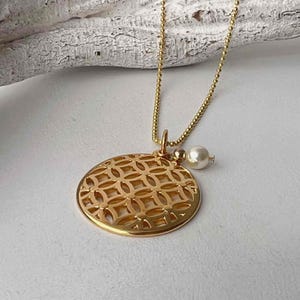 Puede incluir: Un collar dorado con un colgante redondo calado con un patr&oacute;n geom&eacute;trico repetitivo. Un peque&ntilde;o dije de perla cuelga de la cadena. El collar se muestra sobre una superficie blanca.